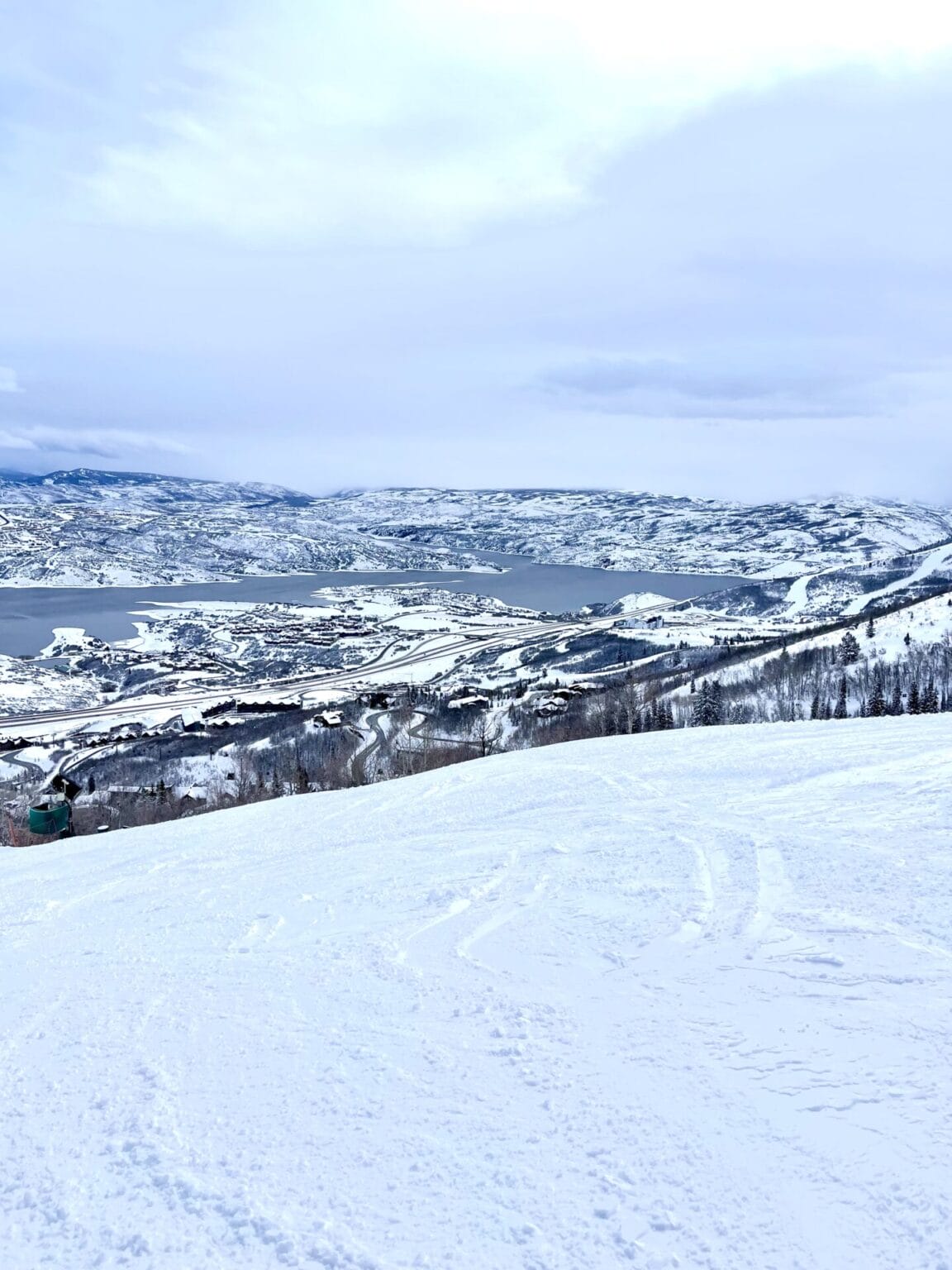 A scenic winter view from a snowy slope overlooking Deer Valley's East Village. The landscape is covered in a thick blanket of snow with patterns from ski activity visible in the foreground. A chairlift can be seen in the middle distance, and the village is nestled among rolling hills that extend to the frozen lake and snow-covered mountains in the distance. The sky is overcast with soft light filtering through the clouds, casting a serene glow over the chilly, picturesque setting.